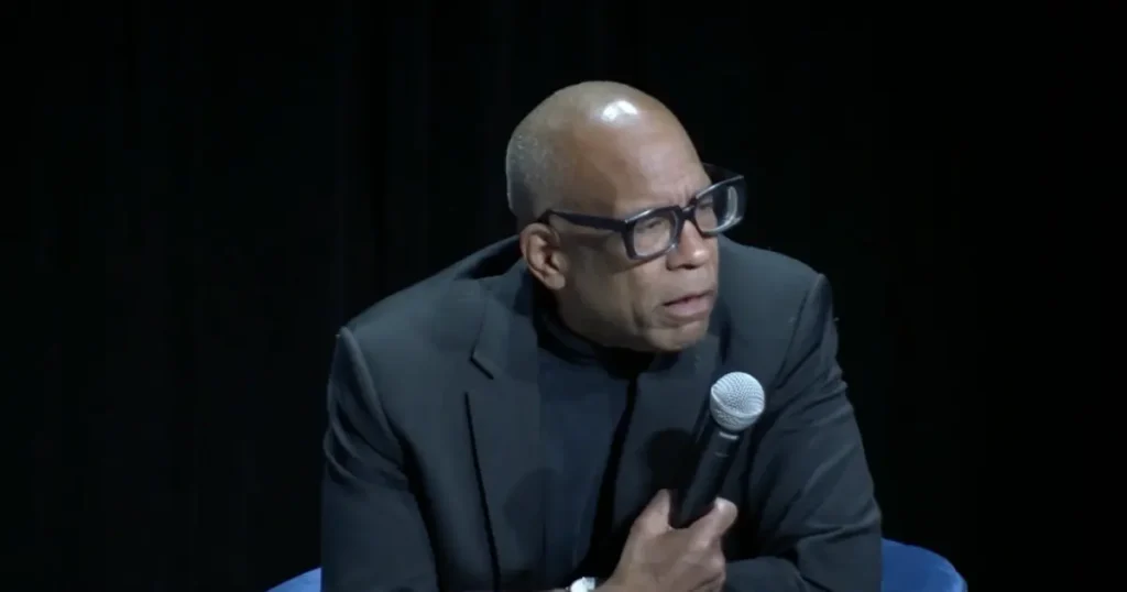 The image shows a person speaking into a microphone, dressed in a dark suit and glasses, against a dark background.