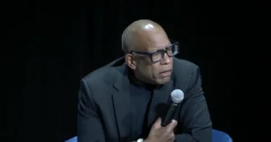 The image shows a person speaking into a microphone, dressed in a dark suit and glasses, against a dark background.