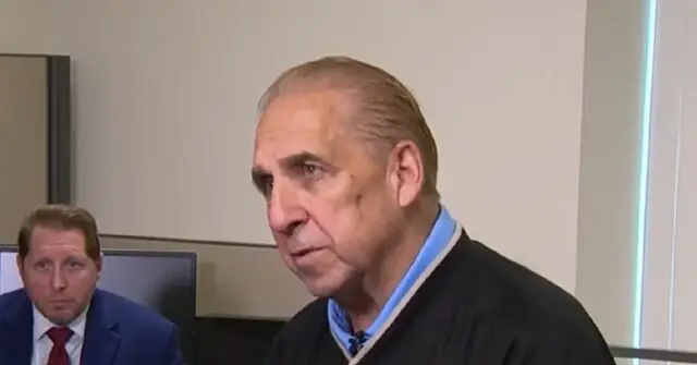 The image shows a man in a judge's robe speaking, with another individual in a suit seated in the background.