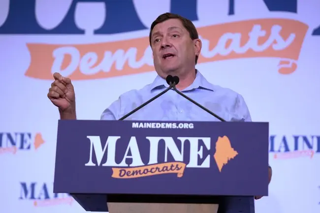 A man stands at a podium with a "Maine Democrats" sign, addressing an audience at a political event.