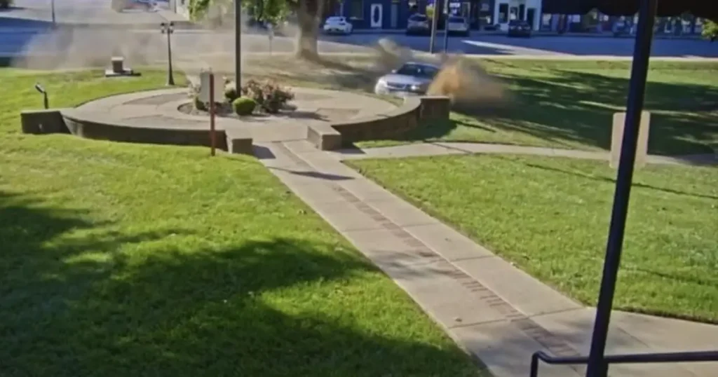 A car is seen driving through a landscaped area, causing a cloud of dust to rise.