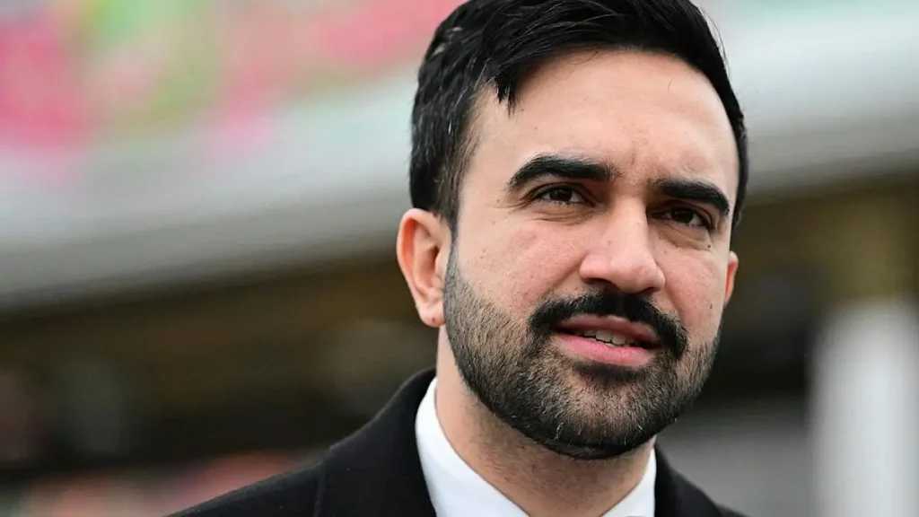 The image shows a man with a beard and short hair, dressed in a suit, standing in an outdoor setting with a blurred background.