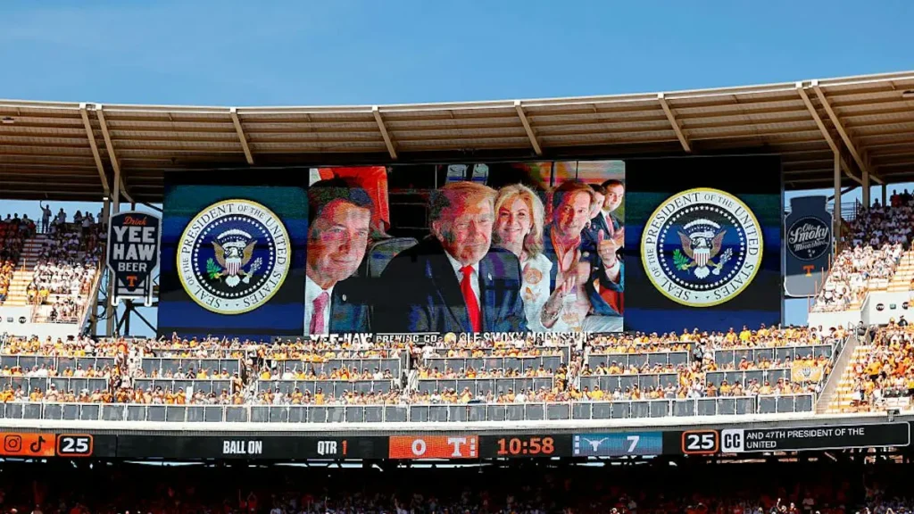 The image shows a large stadium screen displaying the Presidential Seal and a group of individuals, with a crowd of spectators in the stands.