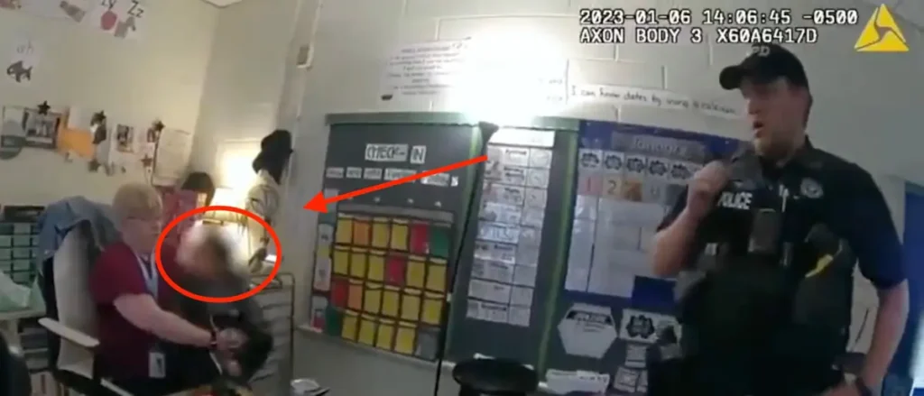 A police officer is seen in a classroom, with a red arrow pointing to a student who appears to be being restrained by another individual.