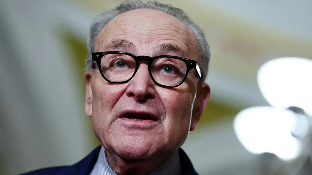 The image shows a close-up of a man wearing glasses and a suit, speaking in what appears to be a formal or professional setting.