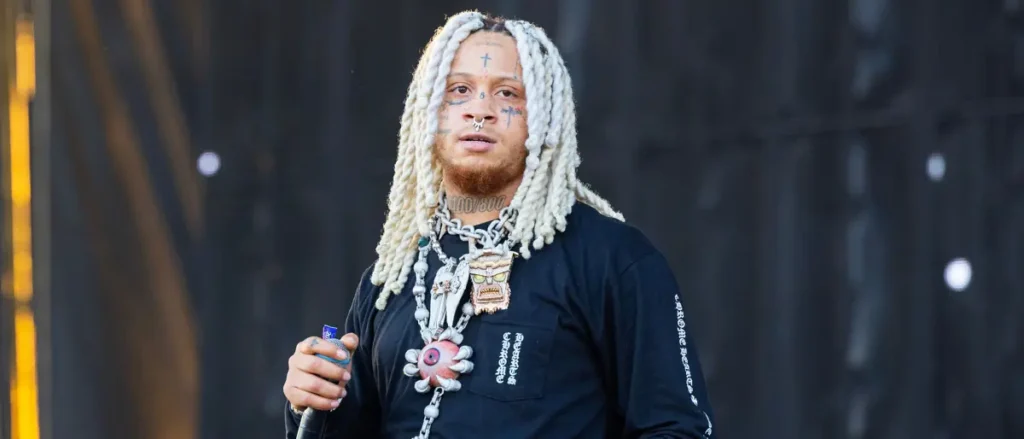 The image shows a person with long, white dreadlocks, wearing a black shirt with a pocket and a chain necklace, holding a microphone, and standing on a stage.