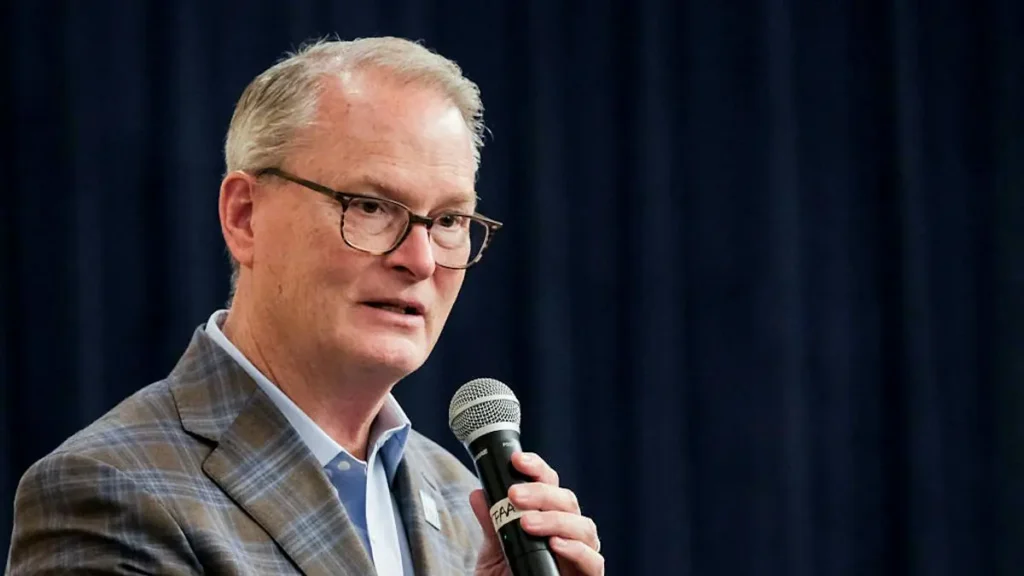 The image shows a man speaking into a microphone, dressed in a plaid blazer and glasses, against a dark blue backdrop.