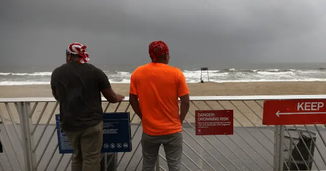 Two individuals stand on a beach observation deck, looking out at the stormy sea, with a warning sign about the drowning risk visible nearby.