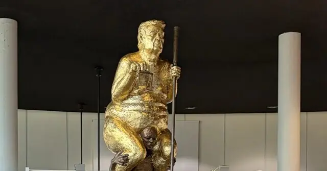 The image shows a controversial statue depicting a figure holding a scale and a stick, standing over another figure, symbolizing power and authority.