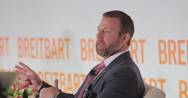 A man in a suit is speaking at a podium with the word "Breitbart" repeated in the background.