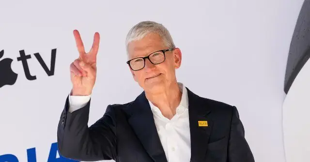 The image shows a person in a suit making a peace sign in front of a backdrop featuring the Apple TV logo.