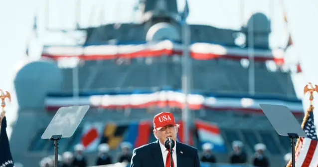 The image shows a person speaking at a podium in front of a large naval ship, with American flags and military personnel in the background.