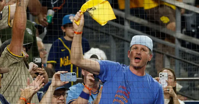 A fan in a blue shirt cheers enthusiastically at a baseball game, holding up a yellow item.