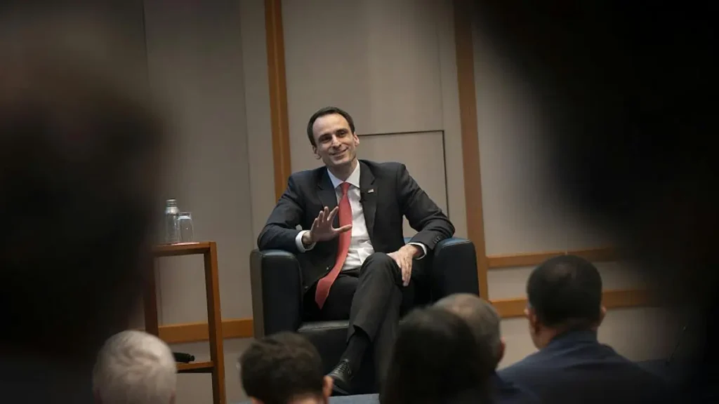 A man in a suit and tie is seated in a chair, engaging with an audience in a formal setting.