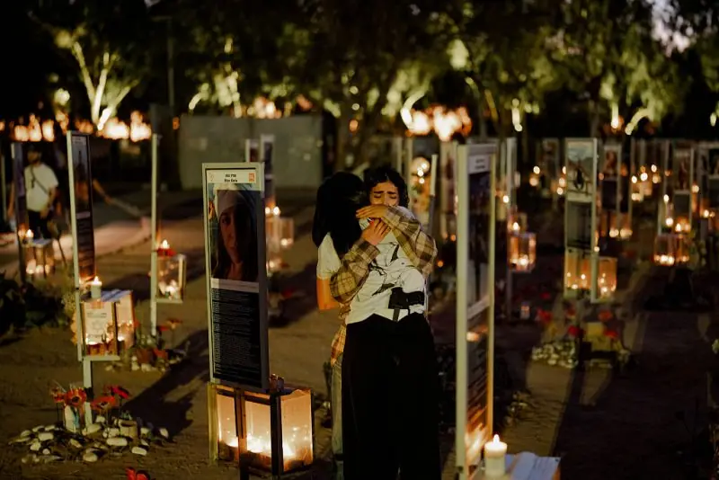 Two individuals embrace in a somber moment at a memorial site adorned with candles and photographs.