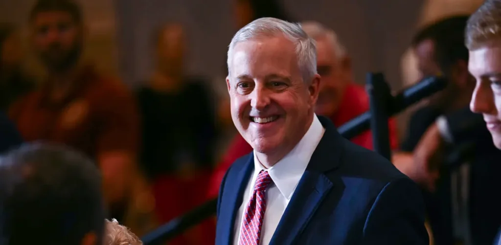 A man in a suit and tie smiles at the camera in a crowded setting.