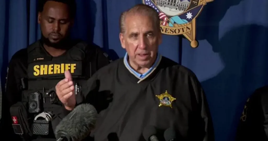 A sheriff speaks at a press conference, with a police officer standing nearby, in front of a Minnesota state emblem backdrop.