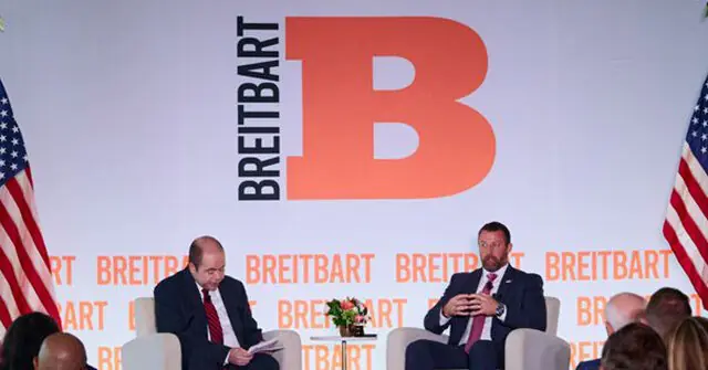 Two men are seated on a stage with a backdrop featuring the "Breitbart" logo and American flags, suggesting a political or media event.