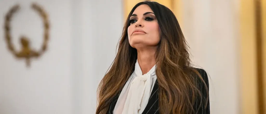 The image shows a person in formal attire, including a white blouse and a dark blazer, standing in a room with a decorative emblem in the background.