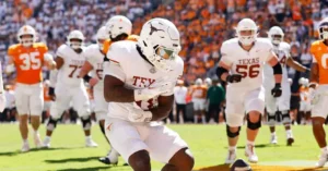 A Texas football player runs onto the field during a game, with teammates and a crowd in the background.