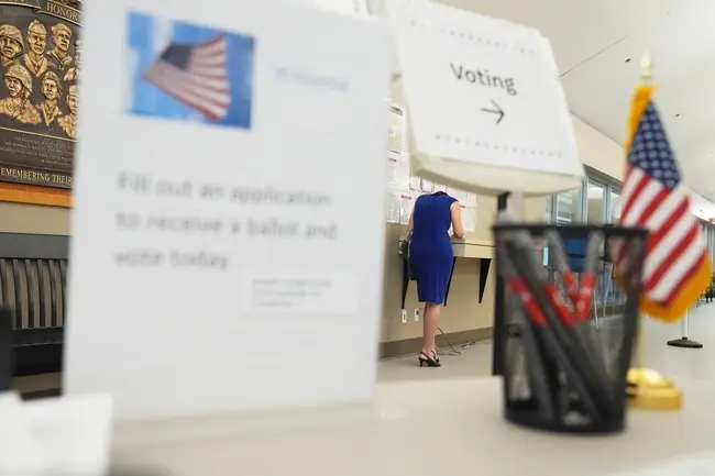 The image depicts a voting station with an American flag, a sign directing voters to the voting area, and a woman in a blue dress filling out an application to receive a ballot.