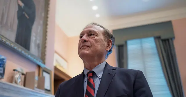 The image shows a man in a suit and tie, looking upward in a formal setting with artwork and a window in the background.