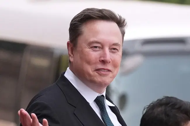 The image shows a man in a suit and tie, gesturing with his hand, with a blurred background that appears to include a vehicle.