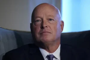 The image shows a man in a suit and tie, seated and looking off to the side with a serious expression.