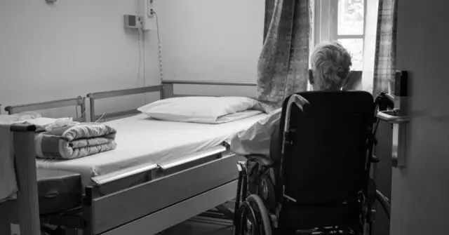 The image depicts a somber scene in a hospital room, featuring an empty bed and a wheelchair, with a person sitting in the wheelchair looking out of a window.