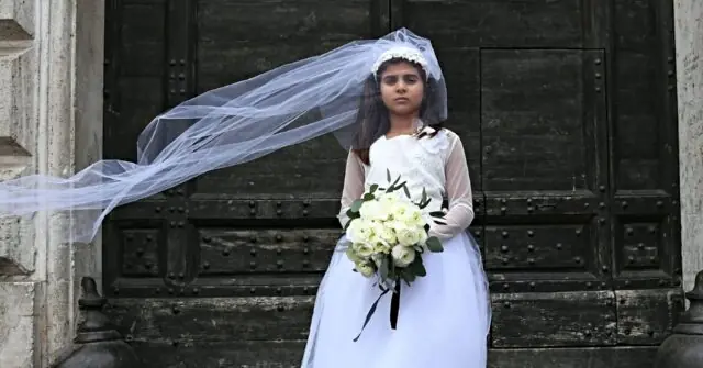 A young girl in a white dress and veil holds a bouquet of flowers, standing in front of a large, ornate door.