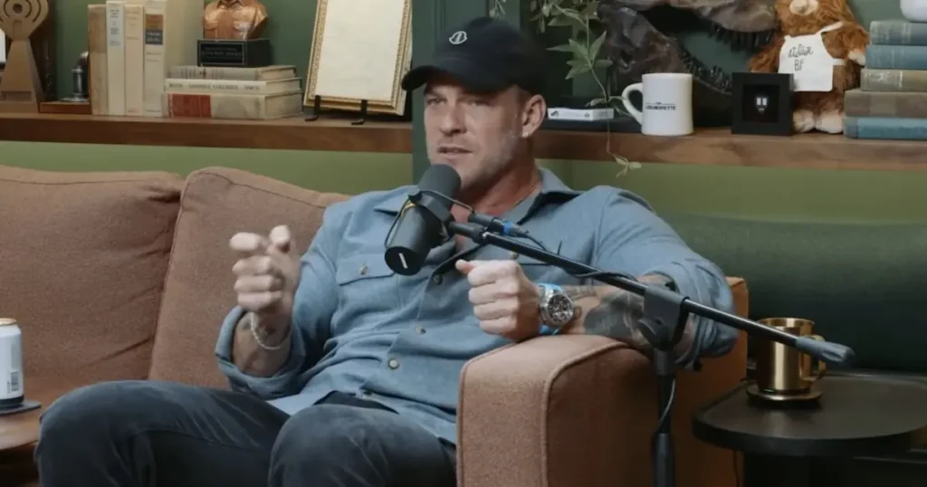 A man is sitting on a couch, speaking into a microphone in a cozy, book-lined room.