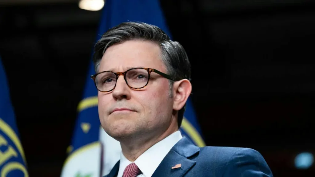 The image shows a man in a suit and tie, wearing glasses, standing in front of a backdrop with flags, suggesting a formal or official setting.