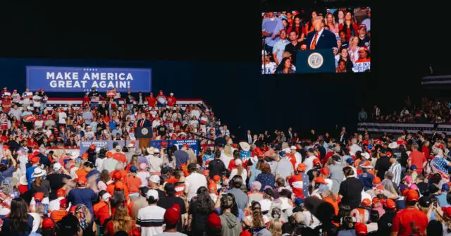 The image depicts a large rally with a crowd gathered, many wearing red hats, and a stage featuring a speaker and a large screen displaying the same scene.
