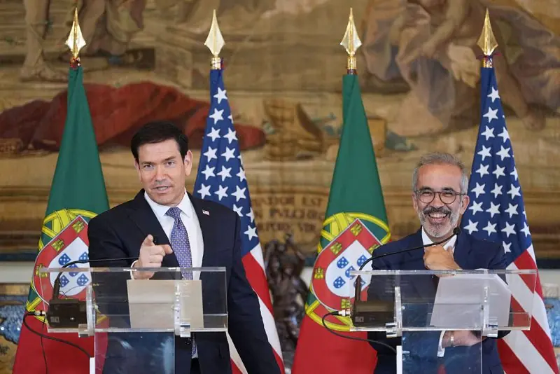 Two individuals are giving speeches at podiums with flags of the United States and Portugal in the background.