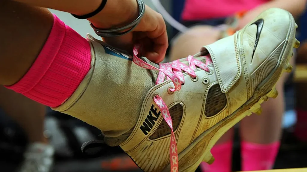 A person is tying the laces of a Nike cleat, which has pink ribbon laces, possibly symbolizing support for breast cancer awareness.