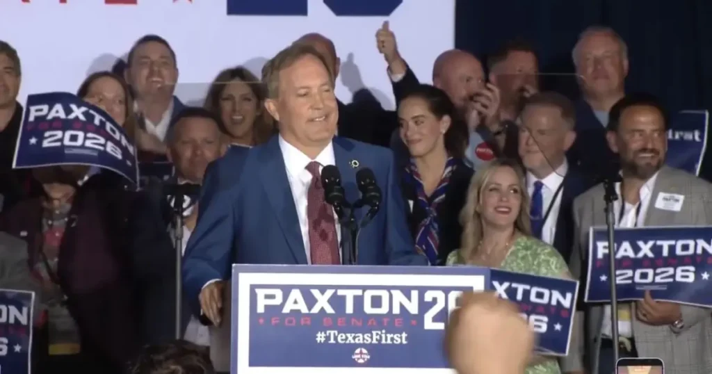 A politician stands at a podium with supporters behind him, holding signs that read "PAXTON 2026" and "TexasFirst."