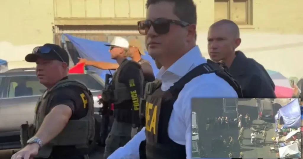The image shows law enforcement officers in tactical gear at a protest or demonstration, with tents and vehicles in the background.
