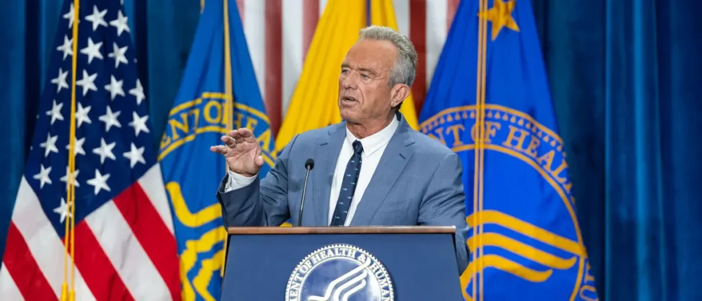 A man in a suit stands at a podium with the Department of Health and Human Services seal, delivering a speech in front of American and Department flags.