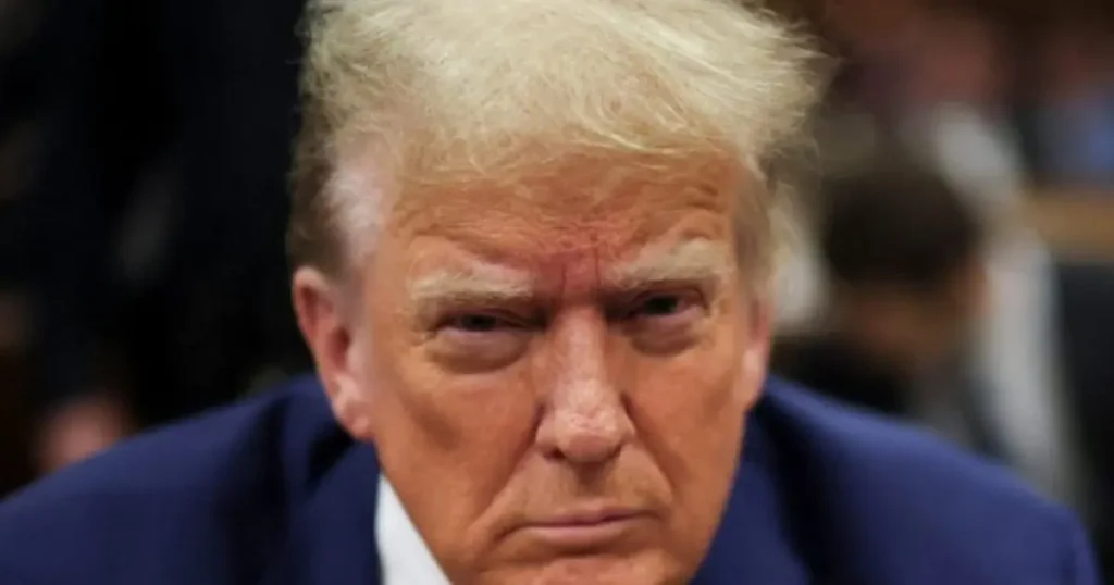 The image shows a close-up of a person in a formal setting, wearing a dark suit and tie, with a serious expression.