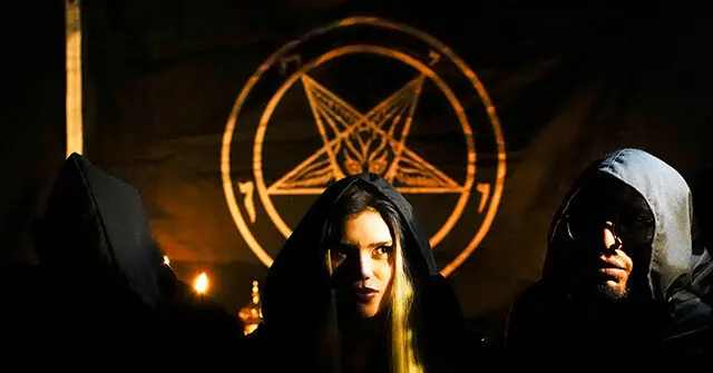 The image depicts a group of hooded individuals standing in front of a pentagram symbol, suggesting a scene from a film or theatrical production involving supernatural or occult themes.