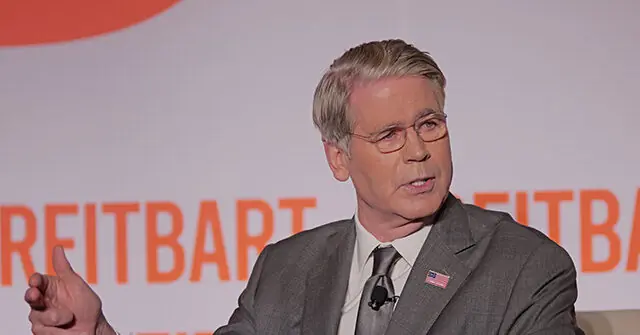The image shows a man in a suit and tie speaking at a podium with the word "REITBART" visible in the background.