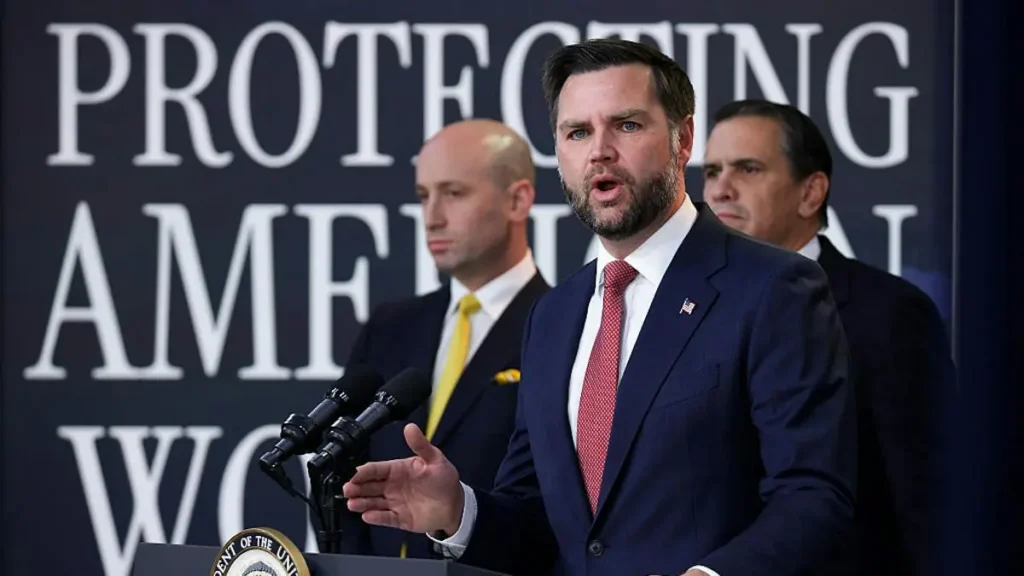A man in a suit stands at a podium with microphones, speaking in front of a backdrop that reads "PROTECTING AMERICA'S WORKERS."