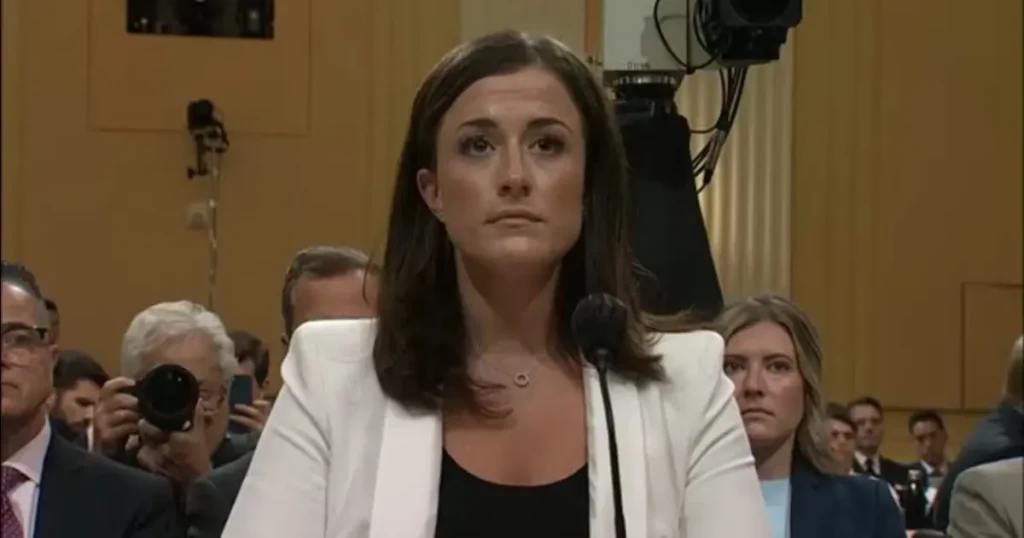 A woman in a white blazer speaks at a formal event, with a microphone in front of her and an audience in the background.
