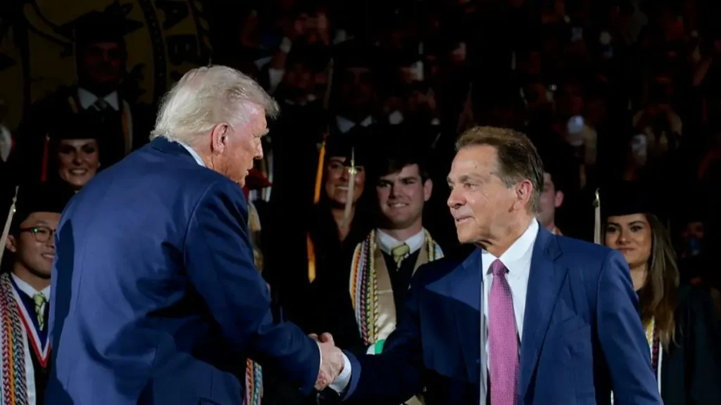 Two men in suits shake hands in front of a crowd of graduates at a formal event.