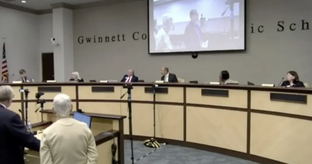The image shows a public meeting or hearing taking place in a conference room with a panel of officials seated at a long table, and a person standing at a podium addressing the panel.