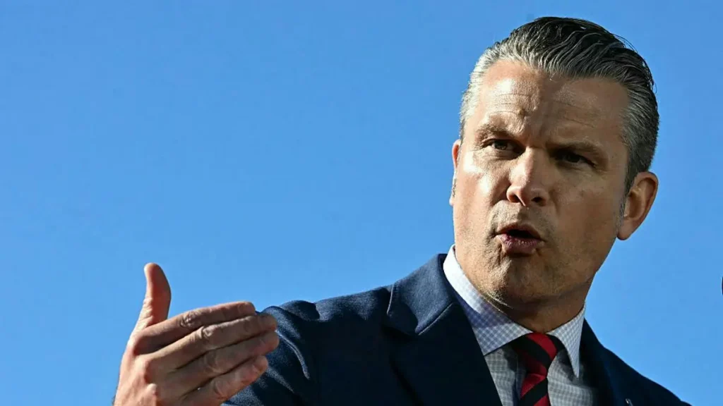The image shows a man in a suit and tie gesturing with his hand against a clear blue sky.