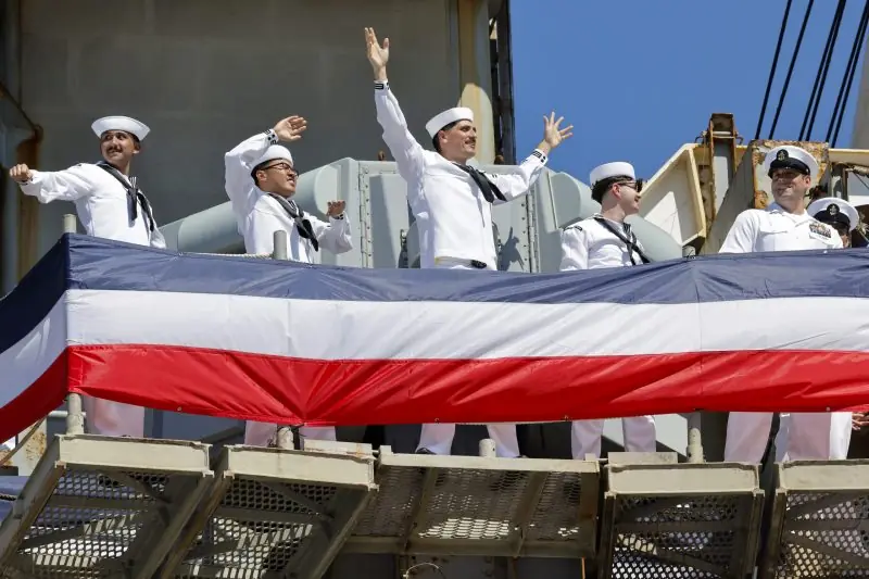 A group of sailors wave and smile from a ship deck adorned with a large American flag.