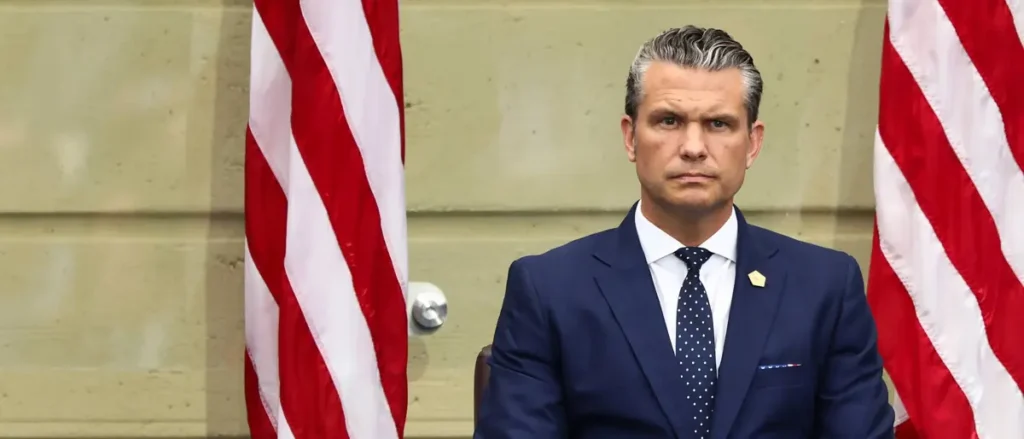 A man in a suit and tie sits in front of two American flags.