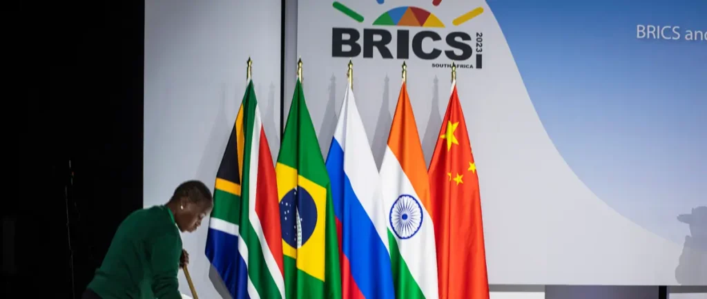 A woman is seen adjusting flags in front of a backdrop featuring the BRICS 2023 logo and the flags of Brazil, Russia, India, China, and South Africa.
