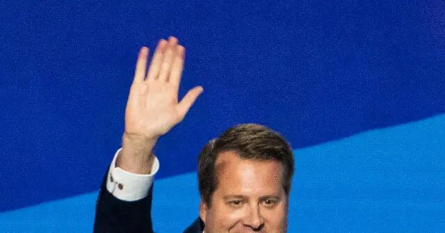 A man in a suit raises his hand in a gesture of greeting or acknowledgment against a blue background.